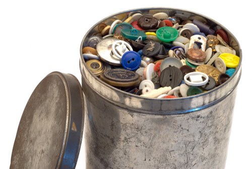 A round metal container filled with a diverse assortment of buttons of various sizes, shapes, and colors, including blue, black, white, green, yellow, and brown, with some metal and plastic finishes visible. The container is situated outdoors on a paved surface near a garden or street setting, with its lid placed to the side. The buttons appear to be discarded or collected items, indicative of waste or rubbish suitable for removal services. The background is plain white or overexposed, focusing attention entirely on the container and its contents, which are relevant to rubbish collection or clearance activities in Osterley and surrounding areas.