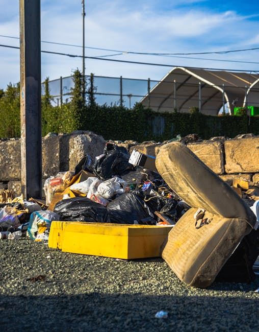 A polluted shoreline scene showing a mixture of plastic waste, including bottles in green, brown, and clear colors, scattered among rocks and pebbles along the water's edge. Several larger rocks and s